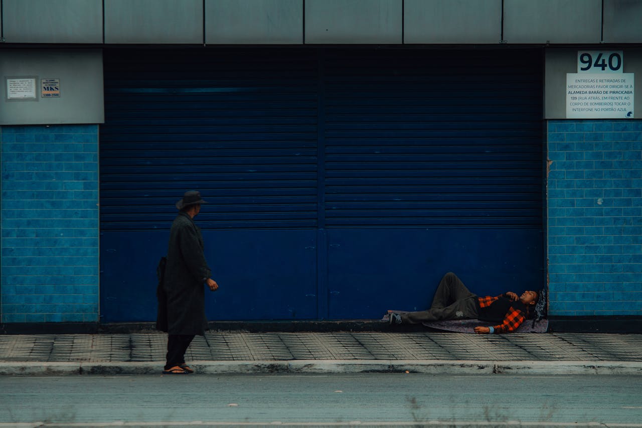 A poignant urban scene depicts a rough sleeper lying on a sidewalk as a passerby walks by.